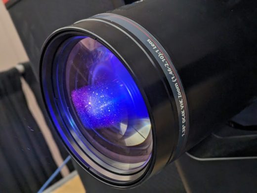Closeup of the lens on a large industrial projector intended for cinema installation displaying an HDR image.