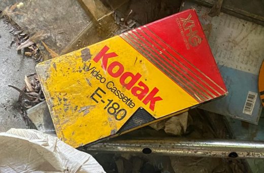 A grubby VHS tape lying in a pile of junk. The tape is branded Kodak in the company's classic red and yellow livery.