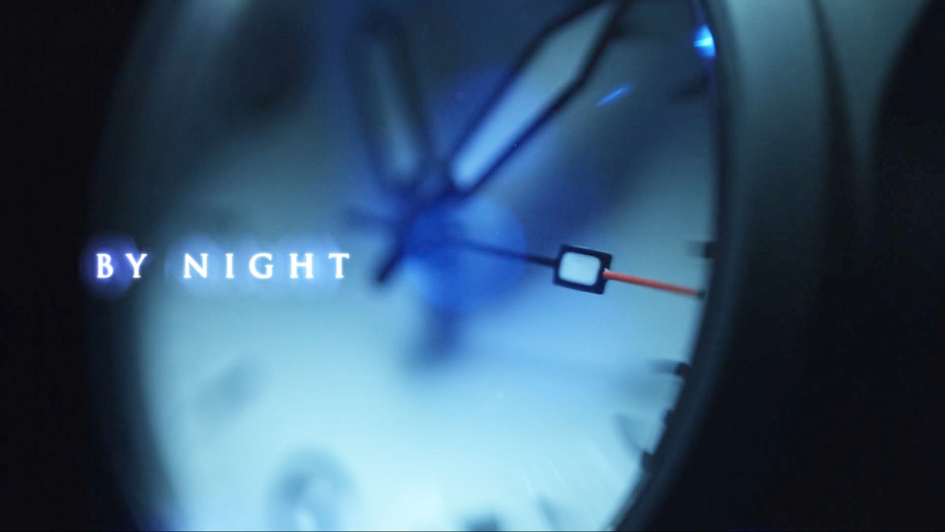 Macro photograph of a watch face against a dark background in shades of cool blue with the text "by night" superimposed.