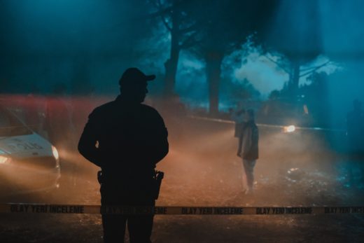 Several people standing in the woods at night behind crime scene tape, a cool blue backlight lancing through mist while the foreground is lit by warmer car headlights.