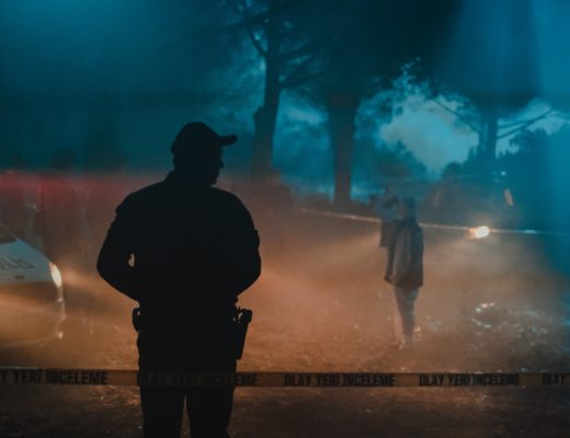 Several people standing in the woods at night behind crime scene tape, a cool blue backlight lancing through mist while the foreground is lit by warmer car headlights.