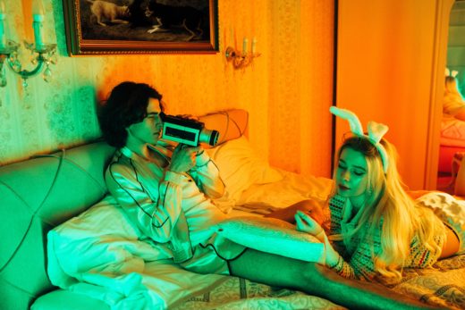 A man lying on a bed films a woman in bunny ears with a super-8mm film camera.