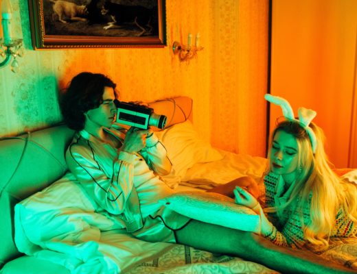 A man lying on a bed films a woman in bunny ears with a super-8mm film camera.