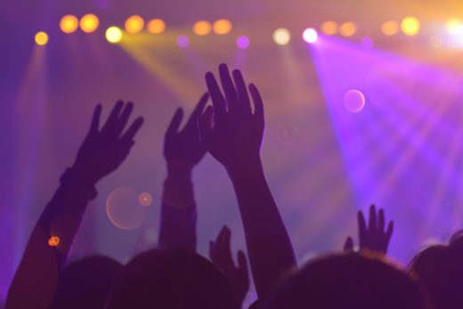 A nightclub crowd dancing with their hands in the air, in front of purple and yellow lights.