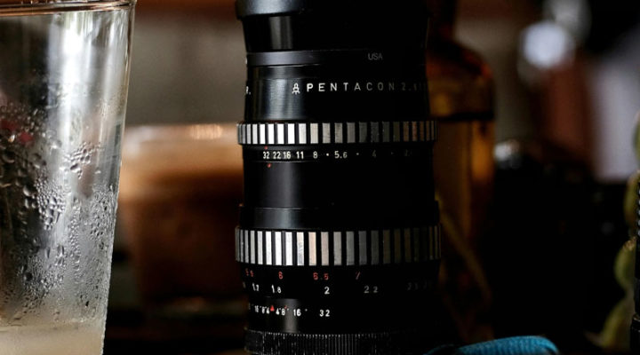 Pentacon lens with zebra-striped barrel on a desk next to a drinking glass.
