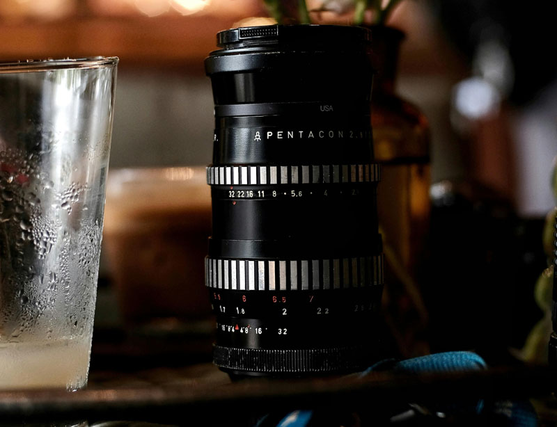 Pentacon lens with zebra-striped barrel on a desk next to a drinking glass.